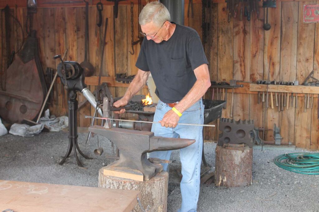 Man in black shirt hammers a hot piece of metal on an anvil in an old time blacksmithing demonstration.