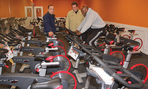 Maximus Health & Fitness owners Frank Distefano (from left) and Phil Durinck look on as personal trainer Greg Trent tries out one of the new LifeCycle GX spin bikes. (Credit: Barbaraellen Koch)