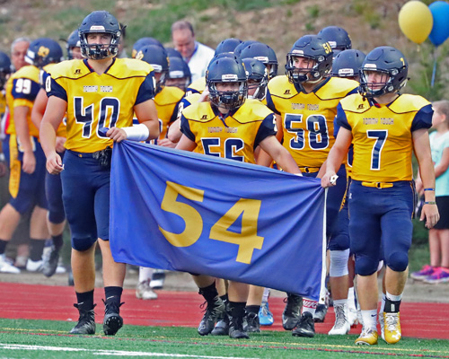 Shoreham-Wading River players Ethan Wiederkehr #40, Logan Snyder #56 and Kevin Cutinella #7 carry the #54 Thomas Cutinella banner onto the field prior to the start of their game against Center Moriches at Shoreham-Wading River High School on September 9, 2016.